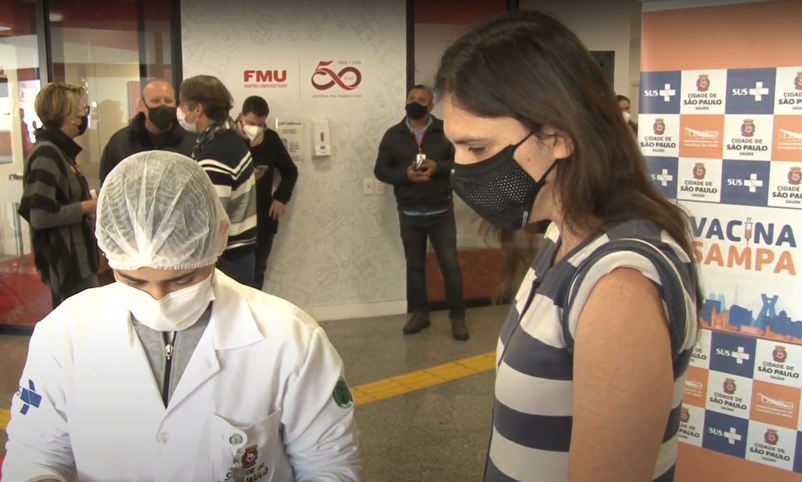 Foto de vacinação. Uma profissional de saúde vestindo um jaleco branco com símbolo do Sistema Único de Saúde (SUS) em uma das mangas, touca de proteção nos cabelos e uma máscara branca está com a cabeça baixa, olhando para um objeto em suas mãos. Ao seu lado, está uma mulher mais alta, com cabelos castanhos, máscara de proteção preta e vestindo uma blusa de listras horizontais brancas e azuis. Uma das mangas está levantada. O painel Vacina Sampa está ao lado direito, nas cores azul, laranja e branco. O logo da faculdade FMU está em uma parede branca ao lado esquerdo, onde também estão algumas pessoas conversando.